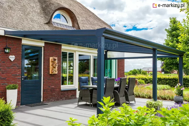 Nowoczesne zadaszenie tarasu model Terano, zamontowane do elewacji budynku wykonanego z czerwonej cegły. Konstrukcja zadaszenia jest aluminiowa, polakierowana na głęboki kolor antracytowy o matowym wykończeniu. Dach ma formę jednospadową o niewielkim kącie nachylenia, a jego wypełnienie stanowią transparentne panele szklane, które pozwalają na swobodny dopływ światła dziennego na taras oraz do wnętrza domu. Stabilność konstrukcji zapewniają pionowe słupy nośne o kwadratowym przekroju, umieszczone na krawędzi tarasu. Widoczny jest zintegrowany system rynnowy w przedniej belce poprzecznej, służący do odprowadzania wody opadowej. Pod zadaszeniem znajduje się strefa wypoczynkowa z dużym stołem obiadowym i sześcioma fotelami z technorattanu z wysokimi oparciami i szarymi poduszkami. Posadzka tarasu wyłożona jest dużymi, jasnoszarymi płytami betonowymi lub kamiennymi. W tle widoczny jest fragment budynku z dachem krytym strzechą oraz zadbany ogród z niską roślinnością i krzewami.