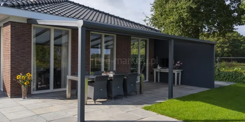 Nowoczesne zadaszene tarasowe Lunaro w kolorze antracytowym, które zostało zamontowane do elewacji parterowego domu wykonanego z czerwonej cegły. Konstrukcja posiada jednospadowy dach wypełniony panelami z poliwęglanu komorowego, wsparty na solidnych, kwadratowych słupach aluminiowych, z których jeden znajduje się na pierwszym planie. Pod zadaszeniem, na jasnych płytach tarasowych, zaaranżowano przestronną jadalnię z dużym stołem o metalowej ramie i sześcioma grafitowymi fotelami z technorattanu. Przy narożniku budynku stoi szklany wazon z bukietem słoneczników, a w głębi, pod pełną ciemną ścianą boczną zadaszenia, ustawiono białą konsolę z kwiatami i dekoracjami. W białych ramach wysokich okien tarasowych odbija się otaczająca zieleń, a całą kompozycję dopełnia widok na idealnie przystrzyżony trawnik, gęstą ścianę drzew w tle oraz dach domu pokryty czarną dachówką.