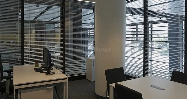 Wnętrze nowoczesnego biura z dużymi przeszkleniami, na których zamocowano system wewnętrznych żaluzji poziomych. Aluminiowe lamele w kolorze srebrnym są ustawione w pozycji otwartej, co pozwala na przenikanie naturalnego światła do pomieszczenia przy jednoczesnym zachowaniu widoczności na sąsiednie strefy biurowe i infrastrukturę zewnętrzną. Pomieszczenie charakteryzuje się surową estetyką z centralnie umieszczonym, okrągłym słupem betonowym oraz ciemną wykładziną podłogową. Wyposażenie obejmuje białe biurka z czarnymi fotelami biurowymi, monitory komputerowe oraz minimalistyczną białą szafkę dokumentową. Przez gęsto rozmieszczone pióra żaluzji widoczne są odbicia konstrukcji sufitu oraz zarysy drzew i budynków za oknem, co podkreśla funkcję systemu w zakresie kontroli nasłonecznienia i prywatności w przestrzeni typu open space.