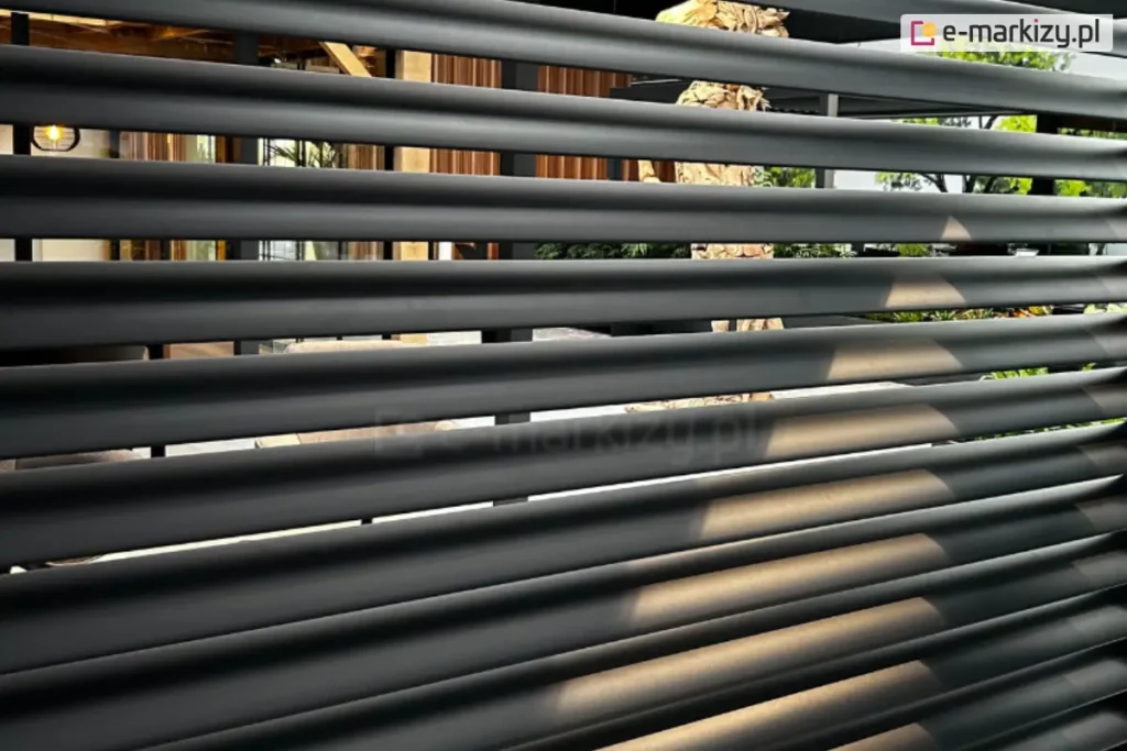 Zdjęcie prezentuje zbliżenie na aluminiowy panel ścienny o konstrukcji żaluzjowej, zamontowany jako boczna przesłona pergoli. Widoczne są poziome, równolegle rozmieszczone lamele o eliptycznym profilu w kolorze ciemnego antracytu, które tworzą nowoczesną strukturę osłonową. Pomiędzy lamelami zachowane są niewielkie prześwity, które pozwalają na ograniczoną widoczność tła, gdzie zarysowuje się wyposażenie tarasu, meble ogrodowe oraz elementy architektury drewnianej. Powierzchnia paneli charakteryzuje się matowym wykończeniem, a padające światło delikatnie podkreśla obły kształt każdej z żaluzji. Konstrukcja stanowi integralną część nowoczesnego systemu zadaszenia, pełniąc funkcję ochrony przed słońcem i zapewniając prywatność użytkownikom przestrzeni wypoczynkowej.
