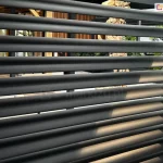 Zdjęcie prezentuje zbliżenie na aluminiowy panel ścienny o konstrukcji żaluzjowej, zamontowany jako boczna przesłona pergoli. Widoczne są poziome, równolegle rozmieszczone lamele o eliptycznym profilu w kolorze ciemnego antracytu, które tworzą nowoczesną strukturę osłonową. Pomiędzy lamelami zachowane są niewielkie prześwity, które pozwalają na ograniczoną widoczność tła, gdzie zarysowuje się wyposażenie tarasu, meble ogrodowe oraz elementy architektury drewnianej. Powierzchnia paneli charakteryzuje się matowym wykończeniem, a padające światło delikatnie podkreśla obły kształt każdej z żaluzji. Konstrukcja stanowi integralną część nowoczesnego systemu zadaszenia, pełniąc funkcję ochrony przed słońcem i zapewniając prywatność użytkownikom przestrzeni wypoczynkowej.