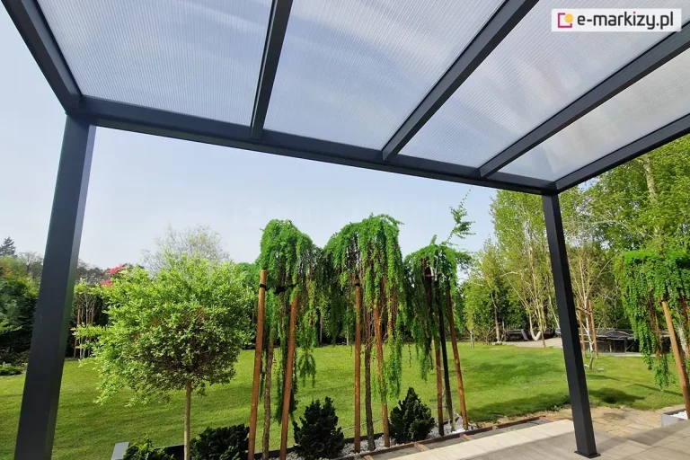 Zdjęcie wykonane z perspektywy żabiej, ukazujące od spodu konstrukcję aluminiowego zadaszenia w kolorze antracytowym. Widoczne są równolegle rozmieszczone krokwie podtrzymujące płyty poliwęglanu komorowego, przez które przebija rozproszone światło dzienne. Konstrukcja opiera się na dwóch skrajnych słupach, tworząc ramę dla widoku na ogród. W tle widać zielony trawnik oraz ozdobne drzewa iglaste o płaczącym pokroju, posadzone w pasie białego żwiru.