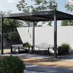 Pergola metalowa Marlo Duo o konstrukcji aluminiowej w kolorze ciemnego grafitu umieszczona w minimalistycznie zaaranżowanym ogrodzie. Konstrukcja pergoli opiera się na czterech solidnych pionowych słupach o kwadratowym przekroju, które podtrzymują płaski dach wyposażony w ruchome lamele pozwalające na regulację dopływu światła słonecznego. Pod zadaszeniem znajduje się zestaw mebli ogrodowych składający się z trzyosobowej sofy, fotela oraz niskiego prostokątnego stolika kawowego. Meble mają czarne ramy i jasnoszare, kontrastujące poduszki siedziskowe oraz oparciowe. Na stoliku widoczne są drobne akcesoria dekoracyjne. Podłoże pod pergolą oraz w jej bezpośrednim sąsiedztwie wykonane jest z dużych, gładkich płyt betonowych w jasnym odcieniu szarości, które przeplatają się z pasami drewnianego podestu o ciepłej barwie. W tle widoczne jest ogrodzenie wykonane z surowego betonu architektonicznego z regularnie rozmieszczonymi otworami technologicznymi, a za nim rosną wysokie liściaste drzewa oraz ozdobne krzewy. Po lewej stronie kompozycji znajdują się dwa przycięte w kulę krzewy posadzone w pasie wysypanym białym grysem, natomiast po prawej stronie widać fragment pionowych drewnianych lameli stanowiących element dekoracyjny ogrodzenia oraz kępy ozdobnych traw. Całość sceny jest oświetlona naturalnym, dziennym światłem, które rzuca wyraźne, geometryczne cienie na nawierzchnię tarasu.