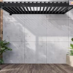 Antracytowa pergola aluminiowa Gloria w wersji przyściennej, zamontowana na nowoczesnym tarasie. Konstrukcja posiada dach z ruchomych lameli, które są ustawione w pozycji otwartej, rzucając geometryczny cień na jasną ścianę budynku wykonaną z betonowych płyt architektonicznych. Po lewej stronie konstrukcji widoczna jest manualna korba służąca do regulacji kąta nachylenia lameli dachowych. Taras jest wyłożony deskami o wyraźnym rysunku drewna, a w jego narożnikach znajdują się minimalistyczne, betonowe donice z gęstą, zieloną roślinnością tropikalną. Tło dla konstrukcji stanowi ściana wykończona kwadratowymi panelami z surowego betonu.