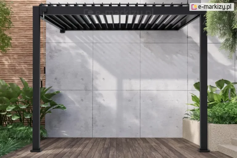 Antracytowa pergola aluminiowa Gloria w wersji przyściennej, zamontowana na nowoczesnym tarasie. Konstrukcja posiada dach z ruchomych lameli, które są ustawione w pozycji otwartej, rzucając geometryczny cień na jasną ścianę budynku wykonaną z betonowych płyt architektonicznych. Po lewej stronie konstrukcji widoczna jest manualna korba służąca do regulacji kąta nachylenia lameli dachowych. Taras jest wyłożony deskami o wyraźnym rysunku drewna, a w jego narożnikach znajdują się minimalistyczne, betonowe donice z gęstą, zieloną roślinnością tropikalną. Tło dla konstrukcji stanowi ściana wykończona kwadratowymi panelami z surowego betonu.