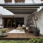 Zdjęcie nowoczesnej pergoli tarasowej o antracytowej, aluminiowej konstrukcji przyściennej, przedstawiona w ujęciu od frontu na tle jasnej elewacji budynku. Zadaszenie składa się z kremowego materiału technicznego, który jest częściowo wysunięty i rozpięty na poprzecznych profilach wyposażonych w zintegrowane, punktowe oświetlenie LED. System dachowy wspiera się na dwóch smukłych, kwadratowych słupach bocznych. Pod pergolą, na tarasie wykonanym z ciemnobrązowych desek kompozytowych o wyraźnym usłojeniu, zaaranżowano strefę wypoczynkową. Znajduje się tam ażurowa sofa i fotel z jasnymi poduchami, drewniany stolik kawowy z bukietem różowych kwiatów oraz biały, prostokątny dywan o grubym splocie. Przestrzeń dekorują rośliny w donicach: po prawej stronie w dużej, grafitowej donicy rośnie ozdobne drzewko o zwisających, bordowych liściach, a po lewej stronie ustawiono dwie obłe, betonowe donice z zielonymi trawami. W tle widoczne są duże, drewniane drzwi tarasowe oraz fragmenty białych ścian bocznych, a u dołu kadru pas zielonego trawnika graniczący z konstrukcją tarasu.