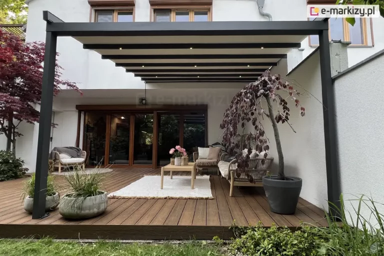 Zdjęcie nowoczesnej pergoli tarasowej o antracytowej, aluminiowej konstrukcji przyściennej, przedstawiona w ujęciu od frontu na tle jasnej elewacji budynku. Zadaszenie składa się z kremowego materiału technicznego, który jest częściowo wysunięty i rozpięty na poprzecznych profilach wyposażonych w zintegrowane, punktowe oświetlenie LED. System dachowy wspiera się na dwóch smukłych, kwadratowych słupach bocznych. Pod pergolą, na tarasie wykonanym z ciemnobrązowych desek kompozytowych o wyraźnym usłojeniu, zaaranżowano strefę wypoczynkową. Znajduje się tam ażurowa sofa i fotel z jasnymi poduchami, drewniany stolik kawowy z bukietem różowych kwiatów oraz biały, prostokątny dywan o grubym splocie. Przestrzeń dekorują rośliny w donicach: po prawej stronie w dużej, grafitowej donicy rośnie ozdobne drzewko o zwisających, bordowych liściach, a po lewej stronie ustawiono dwie obłe, betonowe donice z zielonymi trawami. W tle widoczne są duże, drewniane drzwi tarasowe oraz fragmenty białych ścian bocznych, a u dołu kadru pas zielonego trawnika graniczący z konstrukcją tarasu.