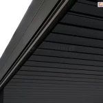 Zbliżenie na narożnik górnej konstrukcji pergoli Gloria w kolorze ciemnoantracytowym, przedstawiające detal prowadnicy i zadaszenia. Zdjęcie wykonane z perspektywy od dołu ukazuje solidną, aluminiową ramę o matowym wykończeniu z precyzyjnie wyprofilowanym wgłębieniem, w którym osadzona jest roleta lub system uszczelniający. Zadaszenie składa się z równolegle ułożonych, płaskich lameli o gładkiej strukturze, które są ciasno dopasowane, tworząc szczelną i jednolitą płaszczyznę w ciemnym odcieniu. W lewym dolnym rogu widoczny jest fragment pionowego słupa nośnego, który łączy się z belką dachową pod kątem prostym, podkreślając nowoczesny i minimalistyczny charakter zabudowy.