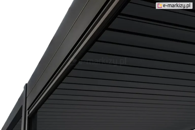 Zbliżenie na narożnik górnej konstrukcji pergoli Gloria w kolorze ciemnoantracytowym, przedstawiające detal prowadnicy i zadaszenia. Zdjęcie wykonane z perspektywy od dołu ukazuje solidną, aluminiową ramę o matowym wykończeniu z precyzyjnie wyprofilowanym wgłębieniem, w którym osadzona jest roleta lub system uszczelniający. Zadaszenie składa się z równolegle ułożonych, płaskich lameli o gładkiej strukturze, które są ciasno dopasowane, tworząc szczelną i jednolitą płaszczyznę w ciemnym odcieniu. W lewym dolnym rogu widoczny jest fragment pionowego słupa nośnego, który łączy się z belką dachową pod kątem prostym, podkreślając nowoczesny i minimalistyczny charakter zabudowy.
