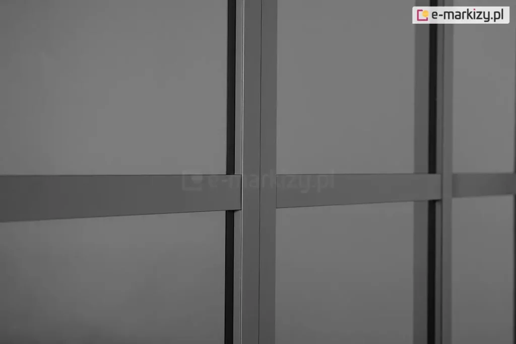 Zbliżenie na detale konstrukcyjne przesuwnych drzwi poliwęglanowych do pergoli Gloria w kolorze ciemnoantracytowym. Zdjęcie koncentruje się na pionowym profilu ramy, który stanowi punkt styku dwóch paneli, oraz na poziomych szprosach wzmacniających, dzielących płaszczyznę drzwi na mniejsze sekcje. Wypełnienie skrzydeł drzwiowych stanowi półprzezroczysty, przydymiony poliwęglan, który delikatnie rozprasza światło, zapewniając prywatność przy jednoczesnym zachowaniu widoczności zarysów otoczenia. Wszystkie elementy aluminiowe charakteryzują się gładkim, matowym wykończeniem i precyzyjnym dopasowaniem krawędzi, co podkreśla nowoczesną estetykę systemu zabudowy.