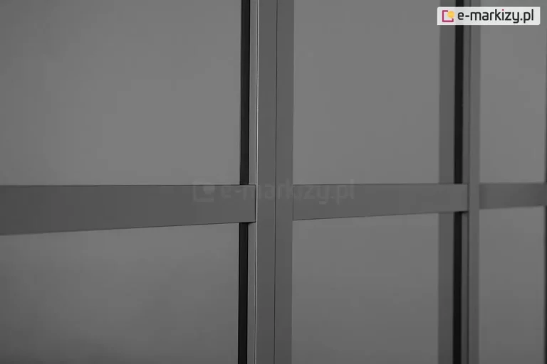 Zbliżenie na detale konstrukcyjne przesuwnych drzwi poliwęglanowych do pergoli Gloria w kolorze ciemnoantracytowym. Zdjęcie koncentruje się na pionowym profilu ramy, który stanowi punkt styku dwóch paneli, oraz na poziomych szprosach wzmacniających, dzielących płaszczyznę drzwi na mniejsze sekcje. Wypełnienie skrzydeł drzwiowych stanowi półprzezroczysty, przydymiony poliwęglan, który delikatnie rozprasza światło, zapewniając prywatność przy jednoczesnym zachowaniu widoczności zarysów otoczenia. Wszystkie elementy aluminiowe charakteryzują się gładkim, matowym wykończeniem i precyzyjnym dopasowaniem krawędzi, co podkreśla nowoczesną estetykę systemu zabudowy.