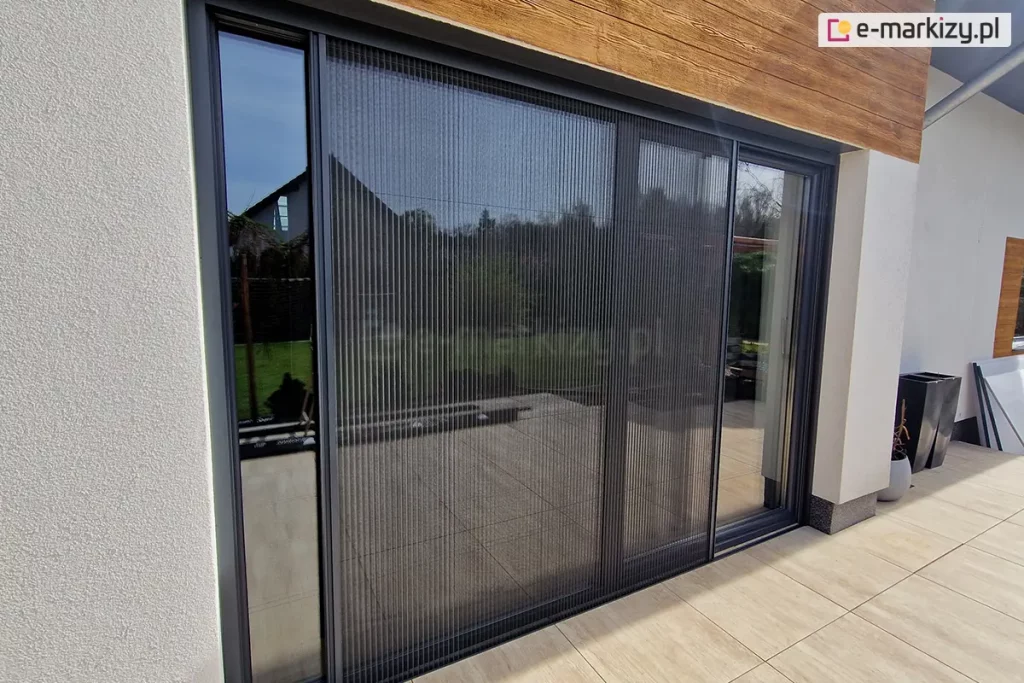Antracytowe drzwi tarasowe przesłonięte częściowo obustronnie otwartą moskitierą plisowaną Top Zag zamocowaną we wnęce o jasnym kolorze elewacji oraz przy tarasie z dużych płytek o jasnym, drewnopodobnym kolorze.