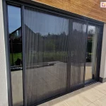 Antracytowe drzwi tarasowe przesłonięte częściowo obustronnie otwartą moskitierą plisowaną Top Zag zamocowaną we wnęce o jasnym kolorze elewacji oraz przy tarasie z dużych płytek o jasnym, drewnopodobnym kolorze.