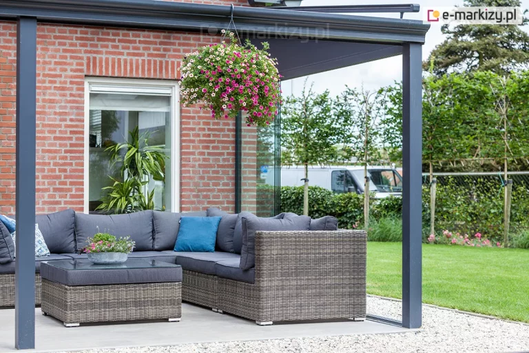 Zdjęcie prezentuje strefę wypoczynkową pod nowoczesnym zadaszeniem tarasowym o konstrukcji aluminiowej w kolorze antracytowym. Pod dachem ustawiono duży narożnik wykonany z technorattanu w odcieniach szarości, wyposażony w dopasowane poduszki siedziskowe i oparciowe oraz jedną dekoracyjną poduszkę w kolorze niebieskim. Przed narożnikiem znajduje się niski stolik kawowy z szklanym blatem. Konstrukcja zadaszenia obejmuje pionowe słupy nośne oraz poziome profile maskujące rynnę, z których zwiesza się donica z obficie kwitnącymi różowo-białymi surfiniami. Boczna część zabudowy została uzupełniona szklanym klinem podkrokwiowym, który oddziela taras od ogrodu, zachowując pełną widoczność. Budynek, do którego przylega konstrukcja, posiada elewację z czerwonej cegły oraz białe ramy okienne, przy których ustawiono wysoką roślinę doniczkową. W tle widoczny jest zadbany trawnik, młode drzewa oraz fragment białego samochodu dostawczego za ogrodzeniem.