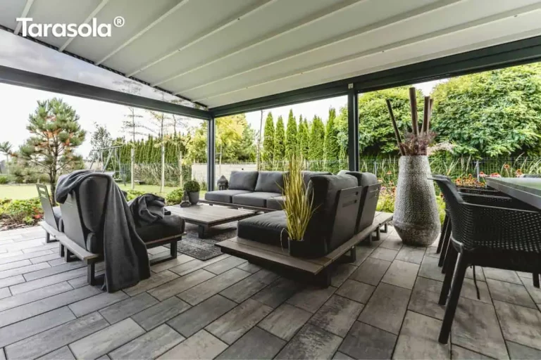 Zdjęcie przedstawia luksusową aranżację dużego tarasu, na którym zainstalowano pergolę tarasola elegancy w wersji z zadaszeniem tkaninowym. Widok z wnętrza konstrukcji ukazuje jasne, segmentowe poszycie dachu rozpięte na antracytowych profilach aluminiowych. Przestrzeń wypoczynkowa została wyposażona w nowoczesne meble ogrodowe z ciemnoszarymi poduszkami na drewnianych podstawach, ustawione na podłożu z szarych płyt tarasowych. W centralnej części znajduje się stolik kawowy oraz dekoracyjne elementy, takie jak wysoki, zdobiony wazon z suchymi pędami i donice z roślinnością. Całość jest otwarta na zadbany ogród z zielonym trawnikiem, gęstym żywopłotem z tui oraz drzewami iglastymi, co podkreśla integrację modelu tarasola elegancy z otoczeniem i jego rolę w tworzeniu komfortowej strefy relaksu.