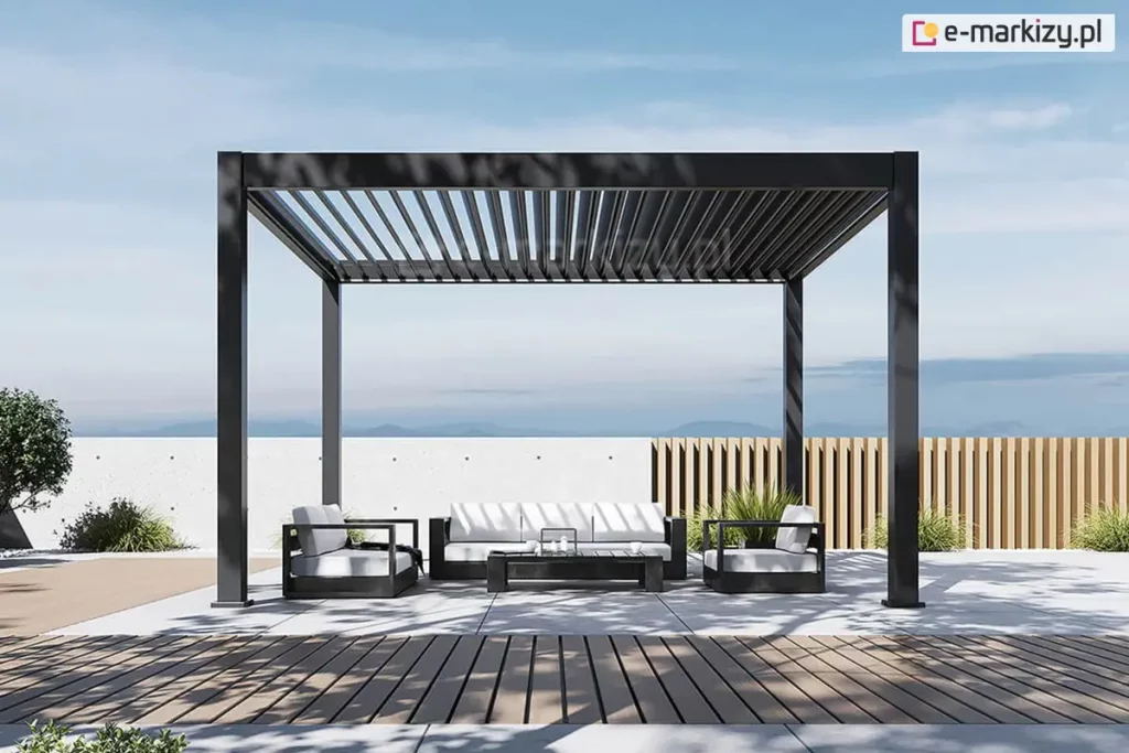 Wolnostojąca pergola bioklimatyczna Rubio w kolorze antracytowym, przedstawiona na otwartym tarasie z widokiem na zamglony horyzont i pasma górskie. Aluminiowy dach lamelowy znajduje się w pozycji otwartej, z lamelami ustawionymi pionowo, co demonstruje funkcję regulacji nasłonecznienia i naturalnej cyrkulacji powietrza. Pod pergolą zaaranżowano nowoczesną strefę wypoczynkową z czarnymi meblami ogrodowymi i białymi poduszkami, ustawionymi na jasnoszarych płytach tarasowych. Po prawej stronie widoczne jest stylowe ogrodzenie z jasnych, pionowych listew drewnianych, a w tle niska, biała ściana z surowego betonu. Nawierzchnia tarasu płynnie łączy się z pasem wykonanym z brązowej deski kompozytowej. Całość kompozycji dopełniają formowane krzewy oraz wysokie trawy ozdobne, a oświetlenie dziennego, bezchmurnego nieba tworzy wyraźne, geometryczne cienie na posadzce tarasu.