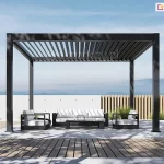 Wolnostojąca pergola bioklimatyczna Rubio w kolorze antracytowym, przedstawiona na otwartym tarasie z widokiem na zamglony horyzont i pasma górskie. Aluminiowy dach lamelowy znajduje się w pozycji otwartej, z lamelami ustawionymi pionowo, co demonstruje funkcję regulacji nasłonecznienia i naturalnej cyrkulacji powietrza. Pod pergolą zaaranżowano nowoczesną strefę wypoczynkową z czarnymi meblami ogrodowymi i białymi poduszkami, ustawionymi na jasnoszarych płytach tarasowych. Po prawej stronie widoczne jest stylowe ogrodzenie z jasnych, pionowych listew drewnianych, a w tle niska, biała ściana z surowego betonu. Nawierzchnia tarasu płynnie łączy się z pasem wykonanym z brązowej deski kompozytowej. Całość kompozycji dopełniają formowane krzewy oraz wysokie trawy ozdobne, a oświetlenie dziennego, bezchmurnego nieba tworzy wyraźne, geometryczne cienie na posadzce tarasu.