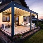 Nowoczesna, przyścienna pergola bioklimatyczna essential Tarasola w kolorze ciemnoantracytowym, zamontowana nad tarasem wykonanym z drobnej kostki brukowej. Konstrukcja posiada dach z ruchomymi lamelami ustawionymi pod kątem oraz zintegrowane oświetlenie LED w formie listew biegnących wzdłuż wewnętrznej krawędzi ramy, które emituje ciepłe światło. Pod zadaszeniem zaaranżowano strefę jadalnianą z białym stołem i krzesłami oraz strefę wypoczynkową z białą sofą i stolikiem kawowym ustawionym na dekoracyjnym dywanie. Pergola przylega do jasnej elewacji domu jednorodzinnego z brązową stolarką okienną, a cała instalacja jest posadowiona na podwyższeniu z ciemnym obrzeżem. Zdjęcie wykonane o zmierzchu ukazuje nastrojową atmosferę ogrodu z widocznym w tle trawnikiem, ciemnym żywopłotem oraz subtelnym, wieczornym niebem w odcieniach fioletu i pomarańczu.