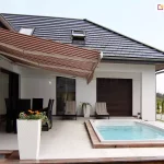 Szeroka markiza tarasowa Palladio w drobne, podłużne pasy łączące odcienie bordowego, beżu i bieli, zamontowana do białej elewacji nowoczesnego budynku mieszkalnego. Konstrukcja z rozłożonym materiałem rzuca wyraźny cień na część wypoczynkową przydomowego tarasu, wyłożonego jasnymi płytami wielkoformatowymi. Pod zadaszeniem usytuowany jest zestaw ciemnych mebli ogrodowych składający się ze stołu i krzeseł, a w głębi podcienia widoczne są dwa leżaki z pasiastą tapicerką, rozdzielone stojącą, białą donicą z zieloną rośliną. Na pierwszym planie, po lewej stronie, stoi wysoka, prostokątna donica o strukturalnej powierzchni z kwitnącymi pomarańczowymi kwiatami. Po prawej stronie kadru znajduje się prostokątny, wbudowany w grunt basen z błękitną wodą, wykończony wokół krawędzi ciemniejszym obrzeżem. Budynek posiada spadzisty dach pokryty grafitową dachówką z widocznym oknem dachowym oraz ciemne ramy okienne i zamknięte rolety zewnętrzne. W tle po prawej stronie majaczy fragment ogrodu z rzędem niskich iglaków oraz podjazd.