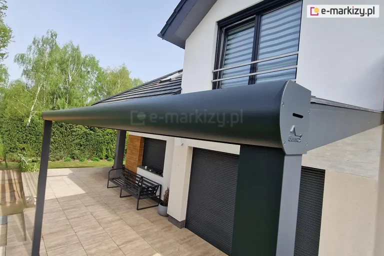 szczegółowe ujęcie konstrukcyjne zakończenia aluminiowego zadaszenia tarasowego w kolorze antracytowym. Na pierwszym planie widoczna jest masywna, zaokrąglona belka czołowa, która pełni funkcję rynny odprowadzającej wodę, zamknięta estetyczną zaślepką boczną z logo producenta. Belka jest zintegrowana z pionowym słupem nośnym o kwadratowym przekroju. Zadaszenie pokryte jest płytami z poliwęglanu, które są zamocowane w aluminiowych profilach dachowych. Konstrukcja przylega do nowoczesnego, piętrowego budynku o jasnej elewacji. W tle widoczny jest taras wyłożony płytkami imitującymi drewno, na którym stoi metalowa ławka, oraz fragment ogrodu z wysokim żywopłotem i drzewami liściastymi. Okna budynku wyposażone są w ciemne rolety zewnętrzne oraz barierki zabezpieczające ze stali nierdzewnej.