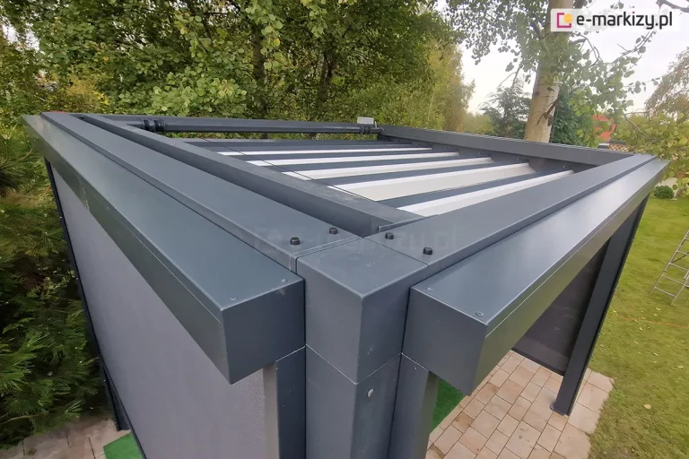 Dwie skrzynki refleksoli zamocowane do ścian bocznych pergoli Tako. Po lewej rozsunięta tkanina rolety w kolorze grafitowym, po prawej widoczne pod pergolą podłoże z kostki brukowej. Dookoła pergoli wiele drzew liściastych.