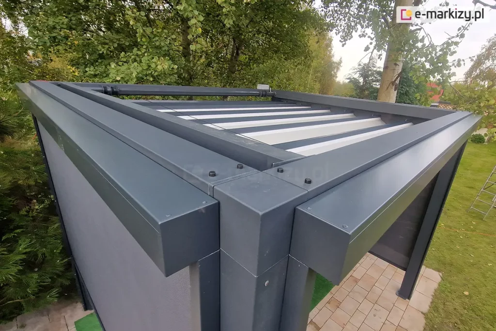 Widok z góry na belki konstrukcji pergoli Tako oraz prowadnice dachu. Po prawej i lewej stronie widoczne kasety rolet screen zamocowanych na dwóch ścianach bocznych. W tle widoczny także rozłożony dach tkaninowy. Pergola ustawiona w ogrodzie, na podłoży wyłożonym kostką brukową w pobliżu zadbanego trawnika, w otoczeniu zielonych drzew.