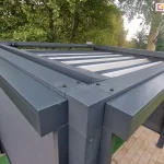 Widok z góry na belki konstrukcji pergoli Tako oraz prowadnice dachu. Po prawej i lewej stronie widoczne kasety rolet screen zamocowanych na dwóch ścianach bocznych. W tle widoczny także rozłożony dach tkaninowy. Pergola ustawiona w ogrodzie, na podłoży wyłożonym kostką brukową w pobliżu zadbanego trawnika, w otoczeniu zielonych drzew.