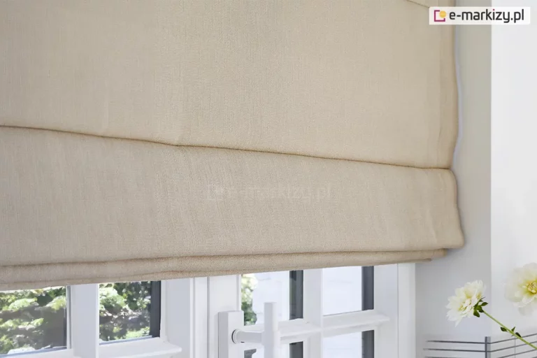 Zdjęcie prezentuje dolną część rolety rzymskiej w kolorze beżowym lub jasnokremowym, zamontowanej bezpośrednio nad białą ramą okienną. Materiał rolety posiada wyraźną, grubszą strukturę splotu, która przypomina naturalną tkaninę typu len lub melonż, z delikatnie widocznymi pionowymi i poziomymi nitkami. Roleta jest częściowo podciągnięta, co tworzy dwie estetyczne, równoległe zakładki materiału nachodzące na siebie poziomo. Dolna krawędź osłony kończy się tuż nad górną linią białych skrzydeł okiennych. Okno pod roletą jest wieloszybowe, z widocznymi pionowymi i poziomymi szprosami dzielącymi przeszklenia na mniejsze pola. W dolnej części kadru widać białą klamkę okienną o prostym, nowoczesnym kształcie. Przez szyby przedostaje się rozproszone światło dzienne, a w tle majaczy zarys zielonej roślinności ogrodowej. Po prawej stronie, na pierwszym planie, widoczny jest fragment białego kwiatu o gęstych płatkach, umieszczonego w wazonie, co ociepla kompozycję i nadaje wnętrzu przytulny, domowy charakter. Ściana otaczająca okno jest gładka i pomalowana na biały kolor, co potęguje wrażenie jasności i przestronności pomieszczenia.