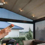 Zbliżenie przedstawia dłoń trzymającą biały pilot zdalnego sterowania z wyświetlaczem, służący do obsługi systemów automatyki w nowoczesnej pergoli tarasola elegancy. W tle widoczne jest wnętrze pergoli z włączonymi punktowymi lampami LED, które są zintegrowane z profilami poprzecznymi jasnego, tkaninowego zadaszenia. Konstrukcja posiada ciemne profile aluminiowe oraz boczne przeszklenia, przez które roztacza się widok na ogród i sąsiedni budynek mieszkalny o jasnej elewacji i niebieskim dachu. Pod zadaszeniem widoczne są fragmenty mebli tarasowych z jasnego technorattanu z ciemnymi poduszkami, co podkreśla komfort i nowoczesny charakter strefy wypoczynkowej. Zdjęcie akcentuje łatwość sterowania oświetleniem i funkcjami pergoli tarasola elegancy za pomocą bezprzewodowych rozwiązań technologicznych.