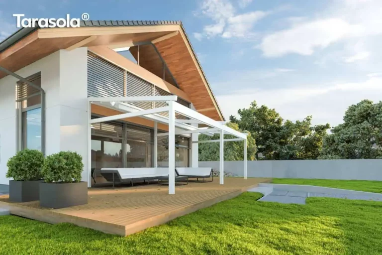 Nowoczesna biała pergola tarasowa Elegancy Tarasola o konstrukcji aluminiowej przyściennej z w pełni złożonym, rzymskim zadaszeniem tkaninowym, zamontowana do elewacji budynku jednorodzinnego. System składa się z wytrzymałych profili nośnych oraz prowadnic dachowych, opartych na trzech pionowych słupach o kwadratowym przekroju. Pod konstrukcją, na przestronnym tarasie wyłożonym naturalnymi deskami drewnianymi, znajduje się duży biały narożnik wypoczynkowy o minimalistycznej formie z ciemnymi stelażami. Budynek charakteryzuje się nowoczesną architekturą z białym tynkiem, dużymi przeszkleniami wyposażonymi w srebrne żaluzje fasadowe oraz szerokim okapem dachowym wykończonym deską w kolorze jasnego drewna. W bezpośrednim otoczeniu tarasu widoczny jest zadbany, zielony trawnik, geometryczne grafitowe donice z formowanymi krzewami oraz pas drzew liściastych w tle pod jasnym, pogodnym niebem.