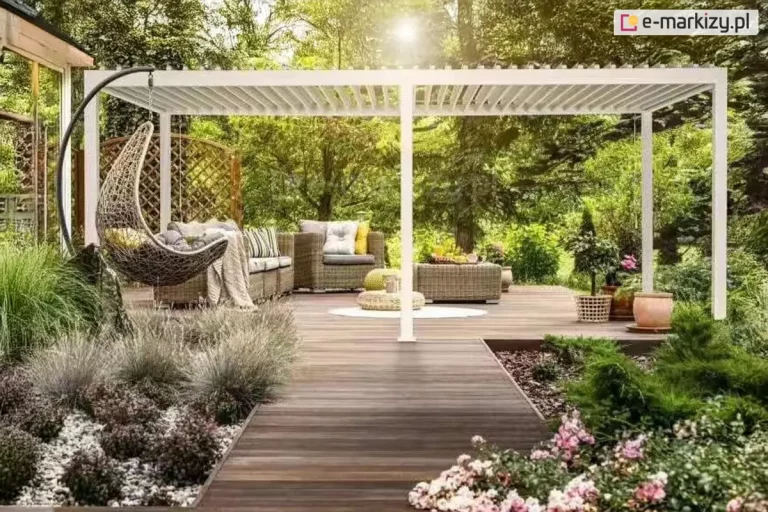 Biała dwumodułowa pergola bioklimatyczna zamontowana na dużym tarasie z ciemnego drewna, wkomponowana w bogato urządzony, słoneczny ogród. Konstrukcja wsparta na solidnych słupach posiada dach z białych, ruchomych lameli ustawionych w pozycji półotwartej. Pod pergolą znajduje się strefa wypoczynkowa z technorattanowymi meblami w kolorze beżowym, obejmująca narożną sofę z poduszkami, fotel oraz niski stolik kawowy, na którym leżą owoce. Po lewej stronie, przed pergolą, zawieszony jest pleciony fotel typu kokon na czarnym stelażu. Taras otoczony jest starannie zaplanowaną roślinnością, w tym trawami ozdobnymi, krzewami iglastymi oraz kwitnącymi na różowo kwiatami w donicach i gruncie. W tle widoczne są wysokie liściaste drzewa, przez które przebijają ostre promienie słońca, tworząc efekt flary. Całość kompozycji prezentuje nowoczesne wykorzystanie pergoli jako funkcjonalnego przedłużenia salonu na świeżym powietrzu.