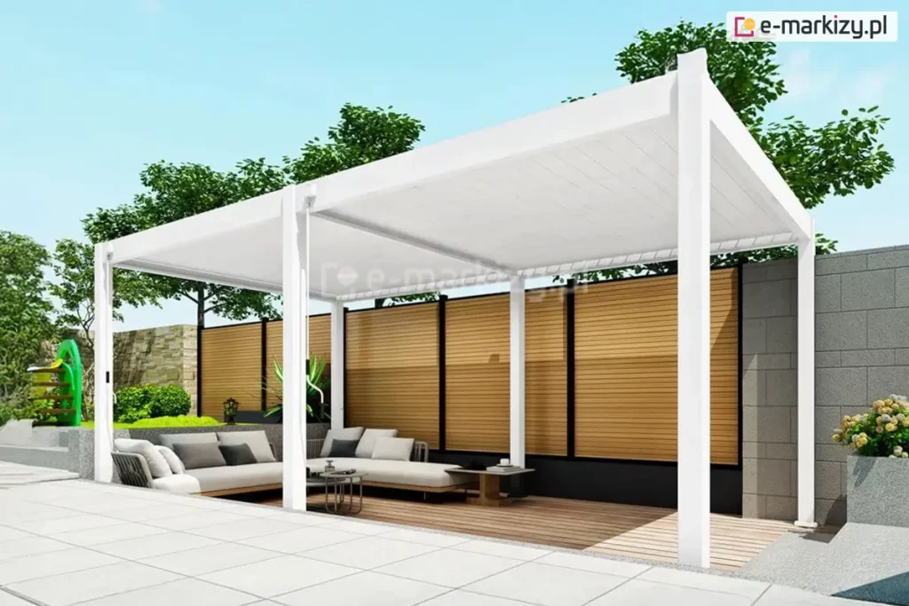 Biała wolnostojąca pergola lamelowa Marlo o szerokości 6 metrów, zamontowana na nowoczesnym tarasie. Konstrukcja składa się z dwóch modułów zadaszenia wspartych na sześciu aluminiowych słupach o kwadratowym przekroju. Ruchome lamele dachowe są całkowicie zamknięte, tworząc szczelną, białą płaszczyznę chroniącą przed słońcem. Pod pergolą, na podłożu z naturalnych desek tarasowych, znajduje się strefa relaksu z dużą jasnoszarą sofą narożną, udekorowaną ciemniejszymi poduszkami, oraz niskie stoliki kawowe o okrągłych blatach. W tle widoczne jest wysokie ogrodzenie wykonane z poziomych drewnianych lameli osadzonych w czarnych ramach, a obok niego betonowy mur z szarych bloczków. Po lewej stronie, w ogrodzie, znajduje się zielony trawnik z elementami placu zabaw dla dzieci oraz gęste korony drzew pod czystym, błękitnym niebem. Na pierwszym planie widoczny jest fragment jasnego, płytowanego tarasu.