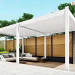 Biała wolnostojąca pergola lamelowa Marlo o szerokości 6 metrów, zamontowana na nowoczesnym tarasie. Konstrukcja składa się z dwóch modułów zadaszenia wspartych na sześciu aluminiowych słupach o kwadratowym przekroju. Ruchome lamele dachowe są całkowicie zamknięte, tworząc szczelną, białą płaszczyznę chroniącą przed słońcem. Pod pergolą, na podłożu z naturalnych desek tarasowych, znajduje się strefa relaksu z dużą jasnoszarą sofą narożną, udekorowaną ciemniejszymi poduszkami, oraz niskie stoliki kawowe o okrągłych blatach. W tle widoczne jest wysokie ogrodzenie wykonane z poziomych drewnianych lameli osadzonych w czarnych ramach, a obok niego betonowy mur z szarych bloczków. Po lewej stronie, w ogrodzie, znajduje się zielony trawnik z elementami placu zabaw dla dzieci oraz gęste korony drzew pod czystym, błękitnym niebem. Na pierwszym planie widoczny jest fragment jasnego, płytowanego tarasu.