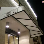 Biała markiza koszowa o sztywnym, trójkątnym profilu i gładkim poszyciu, zamontowana nad wejściem do lokalu usługowego. Na lewym boku konstrukcji widnieje czarny napis LORIBLU, natomiast na frontowym pasie materiału umieszczono napis SPACCIO AZIENDALE wykonany prostą, czarną czcionką. Stelaż markizy składa się z czarnych profili widocznych od spodu, które tworzą geometryczne podziały konstrukcyjne. Osłona jest zainstalowana na jasnej elewacji z piaskowca i oświetlona od góry liniową listwą LED zamocowaną na ścianie oraz od dołu przez dwa punktowe, nowoczesne reflektory sufitowe w kolorze szarym. W tle widoczne są przeszklone drzwi wejściowe oraz fragmenty wnętrza lokalu, a cała scena została uchwycona w warunkach nocnych lub wieczornych.