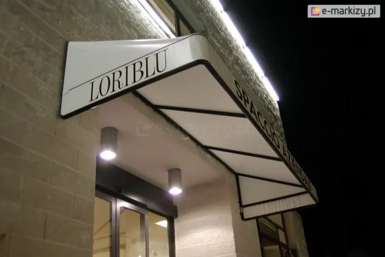 Biała markiza koszowa o sztywnym, trójkątnym profilu i gładkim poszyciu, zamontowana nad wejściem do lokalu usługowego. Na lewym boku konstrukcji widnieje czarny napis LORIBLU, natomiast na frontowym pasie materiału umieszczono napis SPACCIO AZIENDALE wykonany prostą, czarną czcionką. Stelaż markizy składa się z czarnych profili widocznych od spodu, które tworzą geometryczne podziały konstrukcyjne. Osłona jest zainstalowana na jasnej elewacji z piaskowca i oświetlona od góry liniową listwą LED zamocowaną na ścianie oraz od dołu przez dwa punktowe, nowoczesne reflektory sufitowe w kolorze szarym. W tle widoczne są przeszklone drzwi wejściowe oraz fragmenty wnętrza lokalu, a cała scena została uchwycona w warunkach nocnych lub wieczornych.