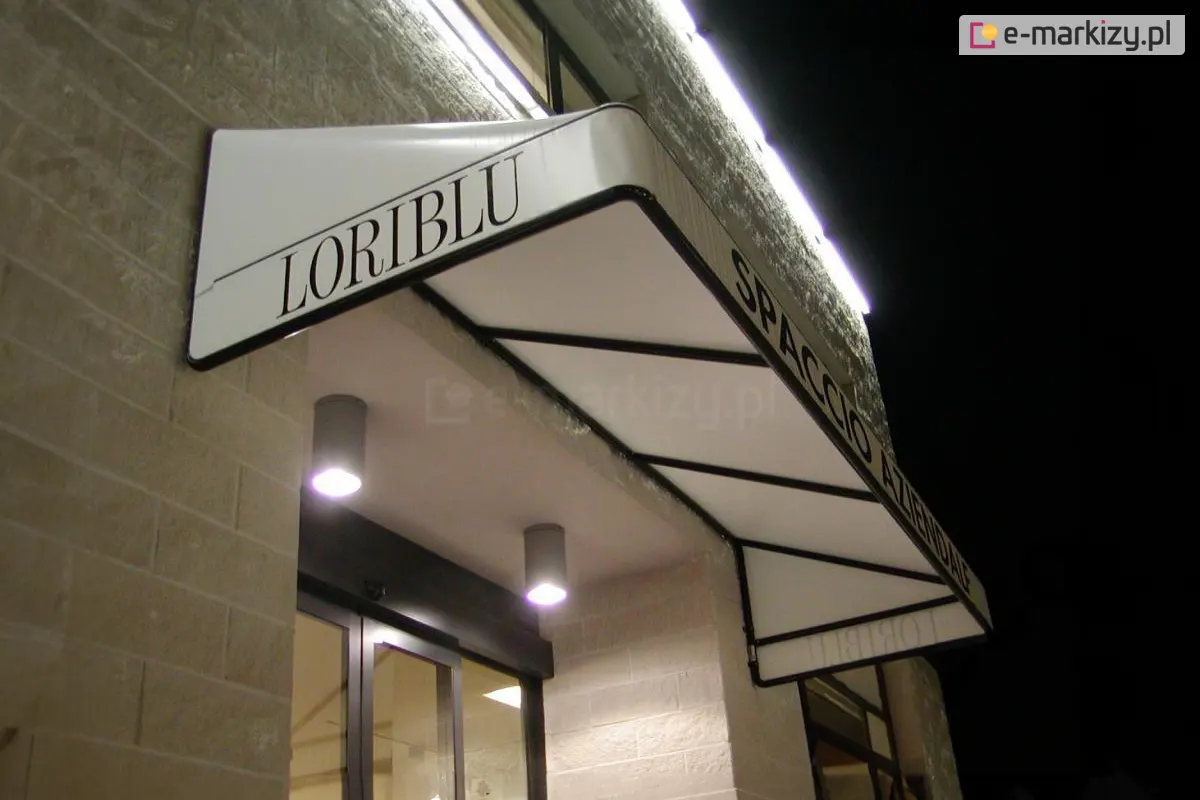 Biała markiza koszowa o sztywnym, trójkątnym profilu i gładkim poszyciu, zamontowana nad wejściem do lokalu usługowego. Na lewym boku konstrukcji widnieje czarny napis LORIBLU, natomiast na frontowym pasie materiału umieszczono napis SPACCIO AZIENDALE wykonany prostą, czarną czcionką. Stelaż markizy składa się z czarnych profili widocznych od spodu, które tworzą geometryczne podziały konstrukcyjne. Osłona jest zainstalowana na jasnej elewacji z piaskowca i oświetlona od góry liniową listwą LED zamocowaną na ścianie oraz od dołu przez dwa punktowe, nowoczesne reflektory sufitowe w kolorze szarym. W tle widoczne są przeszklone drzwi wejściowe oraz fragmenty wnętrza lokalu, a cała scena została uchwycona w warunkach nocnych lub wieczornych.