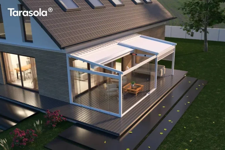 Nowoczesna biała pergola tarasowa o konstrukcji aluminiowej, pełniąca funkcję ogrodu zimowego, zamontowana przy elewacji piętrowego domu jednorodzinnego. Konstrukcja składa się z dwóch modułów wyposażonych w jasne, rozsuwane zadaszenie tkaninowe oraz pełne przeszklenia boczne w systemie przesuwnym, które tworzą zamkniętą przestrzeń rekreacyjną. Wewnątrz obiektu widoczna jest strefa wypoczynkowa z jasnymi sofami, drewnianym stolikiem kawowym oraz roślinnością doniczkową, a pod dachem zamontowano elektryczny promiennik ciepła i zintegrowane oświetlenie punktowe. Budynek charakteryzuje się elewacją z szarymi panelami poziomymi oraz spadzistym dachem z ciemną dachówką i rzędem okien połaciowych. Całość posadowiona jest na tarasie z ciemnych desek kompozytowych, z którego szerokie, czarne stopnie o gładkiej powierzchni prowadzą na zielony trawnik. Zdjęcie wykonane z perspektywy podwyższonej (z lotu ptaka) w scenerii wieczornej przy ciepłym świetle zachodzącego słońca.