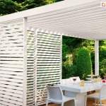 Zbliżenie na narożnik białej pergoli tarasowej wykonanej z aluminium, pod którą urządzono jadalnię w ogrodzie. Konstrukcja posiada zadaszenie z poziomych, białych lameli oraz ściankę boczną wypełnioną stałymi lamelami ułożonymi pod kątem, tworzącymi efekt żaluzji. Pod pergolą ustawiono biały, nowoczesny stół o masywnej podstawie oraz komplet pasujących białych krzeseł z tworzywa. Na stole znajduje się zastawa, szklanki z kolorowymi napojami, kieliszki z winem oraz patera z przekąskami. Tło stanowi intensywnie zielony, gęsty ogród z różnorodnymi krzewami, drzewami liściastymi oraz iglakami, a na jednym z drzew w oddali zawieszona jest drewniana budka lęgowa. Cała scena jest jasno oświetlona naturalnym światłem dziennym, podkreślającym czystą biel konstrukcji i mebli.