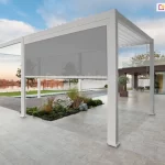 Nowoczesna dwumodułowa pergola bioklimatyczna Gloria w kolorze białym zainstalowana na przestronnym tarasie wyłożonym jasnymi płytami kamiennymi. Konstrukcja z otwartymi lamelami dachowymi współgra kolorystycznie z bryłą nowoczesnego budynku o ciemnej, drewnianej elewacji i dużych przeszkleniach. W jednym z modułów zamontowano szarą roletę screen, która jest częściowo opuszczona, przesłaniając widok na ogród i linię brzegową jeziora w tle. Na tarasie, obok białych słupów pergoli, znajduje się ozdobna wyspa z roślinnością, kamieniami i suchymi trawami. W oddali widoczne są meble ogrodowe ustawione w podcieniu budynku oraz drzewa liściaste podświetlone światłem zachodzącego słońca przy lekko zachmurzonym niebie.