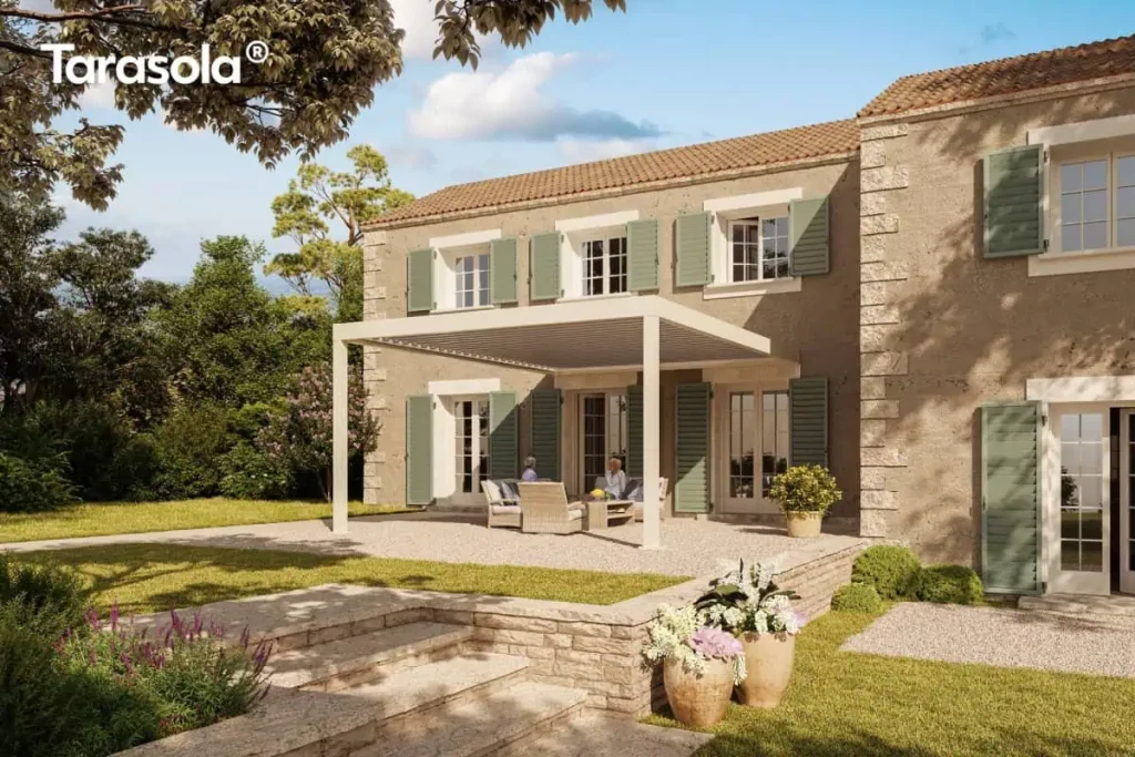 Wizualizacja nowoczesnej pergoli Essential Tarasola w wersji przyściennej, zintegrowanej z architekturą luksusowej rezydencji w stylu śródziemnomorskim. Konstrukcja modelu Essential Tarasola wykonana jest z aluminium w kolorze kremowej bieli i posiada płaski dach lamelowy. Pod zadaszeniem zaaranżowano jasną strefę wypoczynkową z plecionymi meblami ogrodowymi, na którą składają się sofa oraz fotele z szarymi poduszkami, ustawione wokół niskiego stolika kawowego. Budynek mieszkalny o piaszczystej, kamiennej elewacji wyposażony jest w liczne okna i drzwi tarasowe z charakterystycznymi, błękitno-szarymi okiennicami. Cała posesja jest wielopoziomowa; pergola Essential Tarasola znajduje się na podwyższonym tarasie wysypanym jasnym żwirem, do którego prowadzą szerokie, kamienne schody z boku. Na pierwszym planie widoczne są duże, ceramiczne donice z kwitnącymi kwiatami oraz zadbany trawnik otoczony niską roślinnością i lawendą. W tle rozciąga się gęsty pas zielonych drzew, a nad całością widoczne jest pogodne, błękitne niebo z delikatnymi chmurami. Zdjęcie podkreśla elegancki i harmonijny charakter konstrukcji Essential Tarasola w otoczeniu klasycznej architektury i naturalnego ogrodu.
