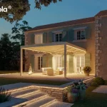 Przestronna pergola bioklimatyczna w kremowo-białym kolorze, z dużym modułem obracanych lameli sterowanych elektrycznie; tworzy elegancką strefę wypoczynkową na tarasie kamiennym przed klasycznym domem z okiennicami, otoczonym bujną zielenią ogrodu.