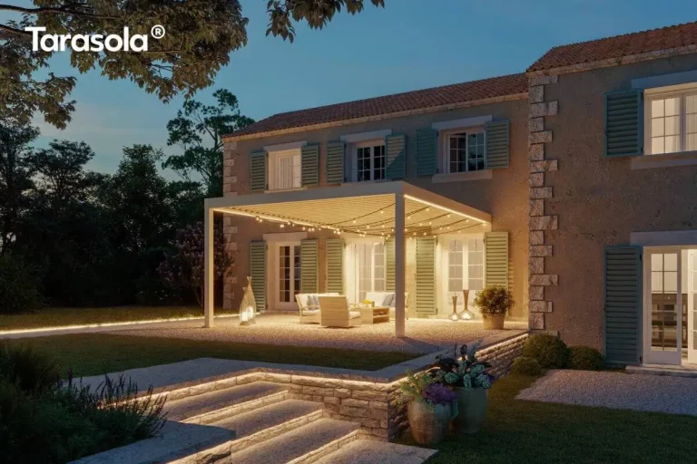 Wieczorna wizualizacja luksusowej rezydencji w stylu śródziemnomorskim z zainstalowaną przyścienną pergolą Essential Tarasola w kolorze białym. Konstrukcja posiada dach lamelowy, pod którym stworzono klimatyczną strefę wypoczynkową, oświetloną zintegrowanymi pasami LED biegnącymi wzdłuż ramy oraz dodatkowymi sznurami dekoracyjnych żarówek podwieszonych pod lamelami. Pod pergolą Essential Tarasola znajdują się jasne meble ogrodowe – sofa oraz fotele z miękkimi poduszkami, ustawione na jasnym, żwirowym podłożu. Budynek charakteryzuje się elewacją z surowego kamienia i tynku w odcieniu ochry, z licznymi oknami i drzwiami balkonowymi ozdobionymi błękitno-szarymi okiennicami. Oświetlenie odgrywa kluczową rolę w tej aranżacji: oprócz iluminacji samej pergoli Essential Tarasola, widoczne są liniowe podświetlenia LED krawędzi kamiennych schodów oraz murków oporowych, a także dekoracyjne lampy stojące w kształcie ażurowych lampionów. Po prawej stronie, na tarasie, umieszczono duże donice z roślinnością, a w tle widać ciemne sylwetki drzew na tle zmierzchającego, błękitnego nieba. Całość aranżacji z modelem Essential Tarasola emanuje spokojem i elegancją, podkreślając luksusowy charakter zewnętrznej strefy relaksu.