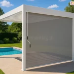 Biała wolnostojąca pergola lamelowa Marlo zamontowana na jasnym tarasie przy basenie, wyposażona w boczna roletę tekstylną typu screen w kolorze beżowo-szarym. Aluminiowa konstrukcja składa się z pionowych słupów o kwadratowym przekroju oraz dachu z ruchomych lamel, które na zdjęciu są ustawione w pozycji zamkniętej. Półprzezroczysta roleta boczna jest całkowicie rozwinięta, przesłaniając jeden z boków pergoli, co zapewnia ochronę przed słońcem przy jednoczesnym zachowaniu widoczności otoczenia. Przy przednim lewym słupie widoczna jest metalowa korba służąca do ręcznej obsługi mechanizmu. Taras wyłożony jest dużymi, kwadratowymi płytami w kolorze piaskowym, a bezpośrednio obok znajduje się basen z błękitną wodą. W tle widoczny jest soczyście zielony, zadbany trawnik oraz gęsta ściana drzew pod pogodnym, błękitnym niebem z nielicznymi białymi chmurami.