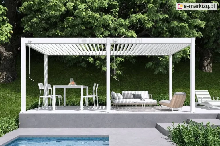 Biała bioklimatyczna pergola tarasowa Marlo o konstrukcji modułowej, zamontowana na podwyższonym betonowym tarasie obok basenu. Zadaszenie składa się z dwóch sekcji ruchomych lamel aluminiowych, które są ustawione pod kątem, wpuszczając rozproszone światło słoneczne na strefę wypoczynkową. Konstrukcja wsparta jest na trzech pionowych słupach frontowych. Po lewej stronie, pod zadaszeniem, znajduje się biały stół jadalniany z czterema pasującymi krzesłami, natomiast po prawej stronie zaaranżowano strefę relaksu z szarą sofą ogrodową, niskim stolikiem kawowym oraz nowoczesnym fotelem z drewnianym wykończeniem. Przy słupach widoczne są mechanizmy korbowe służące do ręcznej regulacji kąta nachylenia lamel. W tle rozpościera się gęsta ściana zielonych drzew, która tworzy naturalne, cieniste tło dla jasnej konstrukcji. Na pierwszym planie widoczny jest fragment krawędzi basenu z błękitną wodą oraz betonowe stopnie prowadzące na taras.