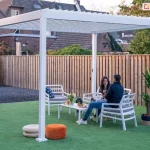 Wolnostojąca bioklimatyczna pergola ogrodowa Gloria w kolorze białym, zamontowana na trawniku w ogrodzie. Konstrukcja oparta na czterech solidnych, kwadratowych profilach aluminiowych z dachem wykonanym z ruchomych lamel ustawionych w pozycji pionowej. Pod pergolą znajduje się biały zestaw mebli ogrodowych: stolik kawowy oraz fotele z szarymi poduszkami, na których siedzi para osób. W aranżacji wykorzystano również dodatki: dwa okrągłe pufy (biały i pomarańczowy) na trawie oraz roślinę doniczkową po prawej stronie. Tło stanowi drewniane ogrodzenie z pionowych desek oraz fragmenty zabudowy mieszkalnej z ceglaną elewacją. Pergola wyposażona jest w manualny mechanizm korbowy widoczny na jednej z tylnych podpór.