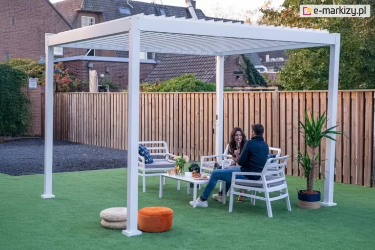 Wolnostojąca bioklimatyczna pergola ogrodowa Gloria w kolorze białym, zamontowana na trawniku w ogrodzie. Konstrukcja oparta na czterech solidnych, kwadratowych profilach aluminiowych z dachem wykonanym z ruchomych lamel ustawionych w pozycji pionowej. Pod pergolą znajduje się biały zestaw mebli ogrodowych: stolik kawowy oraz fotele z szarymi poduszkami, na których siedzi para osób. W aranżacji wykorzystano również dodatki: dwa okrągłe pufy (biały i pomarańczowy) na trawie oraz roślinę doniczkową po prawej stronie. Tło stanowi drewniane ogrodzenie z pionowych desek oraz fragmenty zabudowy mieszkalnej z ceglaną elewacją. Pergola wyposażona jest w manualny mechanizm korbowy widoczny na jednej z tylnych podpór.