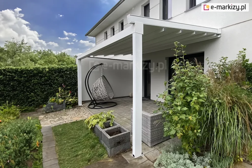 Zdjęcie nowoczesnej pergoli tarasowej o białej, aluminiowej konstrukcji przyściennej, zamontowanej do jasnej elewacji piętrowego budynku. Zadaszenie składa się z białego materiału technicznego rozpiętego na poprzecznych profilach, które w tej konfiguracji są całkowicie wysunięte. Konstrukcja wspiera się na dwóch solidnych, kwadratowych słupach bocznych posadowionych na tarasie wyłożonym szarymi płytami betonowymi. Pod pergolą znajduje się strefa relaksu wyposażona w wiszący fotel typu kosz w kolorze jasnoszarym oraz fragment narożnika z szarego technorattanu. Przestrzeń wokół tarasu jest bogato zagospodarowana roślinnością: po prawej stronie rośnie wysoki krzew o zielonych liściach oraz kępy ozdobnych traw, a po lewej stronie widoczne są betonowe donice z funkiami. Granicę ogrodu wyznacza wysoki, gęsty żywopłot, a w tle widać białą ścianę muru oporowego. Zdjęcie wykonane w dzień, przy błękitnym niebie z delikatnymi białymi chmurami.