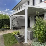 Zdjęcie nowoczesnej pergoli tarasowej o białej, aluminiowej konstrukcji przyściennej, zamontowanej do jasnej elewacji piętrowego budynku. Zadaszenie składa się z białego materiału technicznego rozpiętego na poprzecznych profilach, które w tej konfiguracji są całkowicie wysunięte. Konstrukcja wspiera się na dwóch solidnych, kwadratowych słupach bocznych posadowionych na tarasie wyłożonym szarymi płytami betonowymi. Pod pergolą znajduje się strefa relaksu wyposażona w wiszący fotel typu kosz w kolorze jasnoszarym oraz fragment narożnika z szarego technorattanu. Przestrzeń wokół tarasu jest bogato zagospodarowana roślinnością: po prawej stronie rośnie wysoki krzew o zielonych liściach oraz kępy ozdobnych traw, a po lewej stronie widoczne są betonowe donice z funkiami. Granicę ogrodu wyznacza wysoki, gęsty żywopłot, a w tle widać białą ścianę muru oporowego. Zdjęcie wykonane w dzień, przy błękitnym niebie z delikatnymi białymi chmurami.