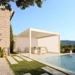 Nowoczesna, biała pergola lamelowa essential Tarasola w wersji wolnostojącej, zainstalowana na jasnym tarasie przy basenie typu infinity. Konstrukcja bioklimatyczna posiada dach z ruchomych lameli oraz zintegrowane boczne rolety materiałowe typu screen, które są w pełni opuszczone na dwóch ścianach, tworząc zamkniętą i osłoniętą przestrzeń wypoczynkową. Pod zadaszeniem widoczny jest zestaw jasnych mebli ogrodowych z miękkimi poduszkami. Pergola znajduje się bezpośrednio przy ścianie budynku z jasnej, dekoracyjnej cegły z szarymi okiennicami. Aranżację uzupełnia wypielęgnowany trawnik z ułożonymi na nim kamiennymi płytami chodnikowymi, niska roślinność w donicach oraz wysoki cyprys. W tle rozpościera się malowniczy widok na zielone wzgórza pod czystym, błękitnym niebem, co podkreśla funkcjonalność i estetykę osłon bocznych w słoneczne dni.