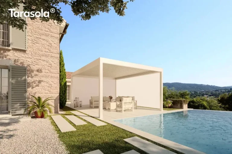 Nowoczesna, biała pergola lamelowa essential Tarasola w wersji wolnostojącej, zainstalowana na jasnym tarasie przy basenie typu infinity. Konstrukcja bioklimatyczna posiada dach z ruchomych lameli oraz zintegrowane boczne rolety materiałowe typu screen, które są w pełni opuszczone na dwóch ścianach, tworząc zamkniętą i osłoniętą przestrzeń wypoczynkową. Pod zadaszeniem widoczny jest zestaw jasnych mebli ogrodowych z miękkimi poduszkami. Pergola znajduje się bezpośrednio przy ścianie budynku z jasnej, dekoracyjnej cegły z szarymi okiennicami. Aranżację uzupełnia wypielęgnowany trawnik z ułożonymi na nim kamiennymi płytami chodnikowymi, niska roślinność w donicach oraz wysoki cyprys. W tle rozpościera się malowniczy widok na zielone wzgórza pod czystym, błękitnym niebem, co podkreśla funkcjonalność i estetykę osłon bocznych w słoneczne dni.